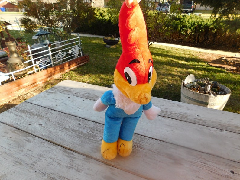 May include: A plush toy of Woody Woodpecker, a cartoon character. The toy is red, yellow, and blue. It is standing on a wooden surface.