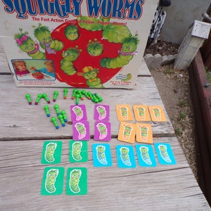 May include: A vintage board game called "Squiggly Worms" with a red apple and green worms on the box. The game includes green worm pieces, cards with worms on them, and a game board.