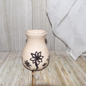 May include: A small, cream-colored ceramic vase with a black and white floral design. The vase has a rounded body and a slightly flared lip.