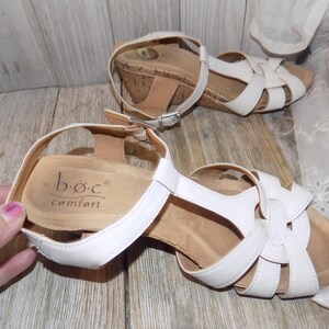 K&ouml;nnte beinhalten: Wei&szlig;e Ledersandalen mit Keilabsatz und Korkplattform sowie Kn&ouml;chelriemen. Die Sandalen haben ein Kreuzdesign und der Markenname "b.o.c. comfort" ist auf der Innensohle aufgedruckt.