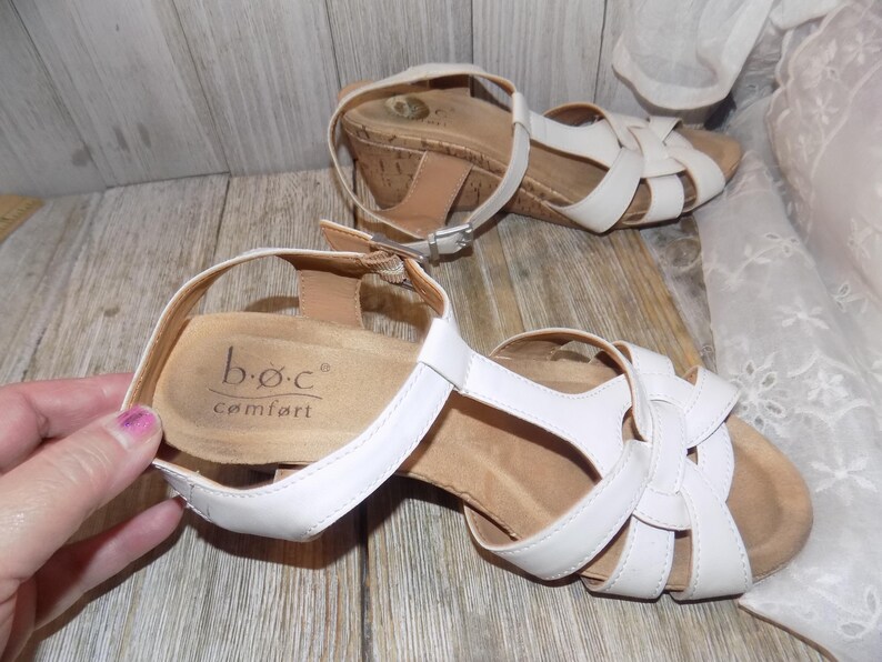 K&ouml;nnte beinhalten: Wei&szlig;e Ledersandalen mit Keilabsatz und Korkplattform sowie Kn&ouml;chelriemen. Die Sandalen haben ein Kreuzdesign und der Markenname "b.o.c. comfort" ist auf der Innensohle aufgedruckt.
