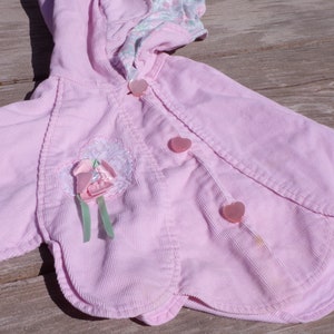 Puede incluir: Una chaqueta de pana rosa con capucha y botones en forma de coraz&oacute;n. La chaqueta tiene un adorno de flor en la parte delantera.
