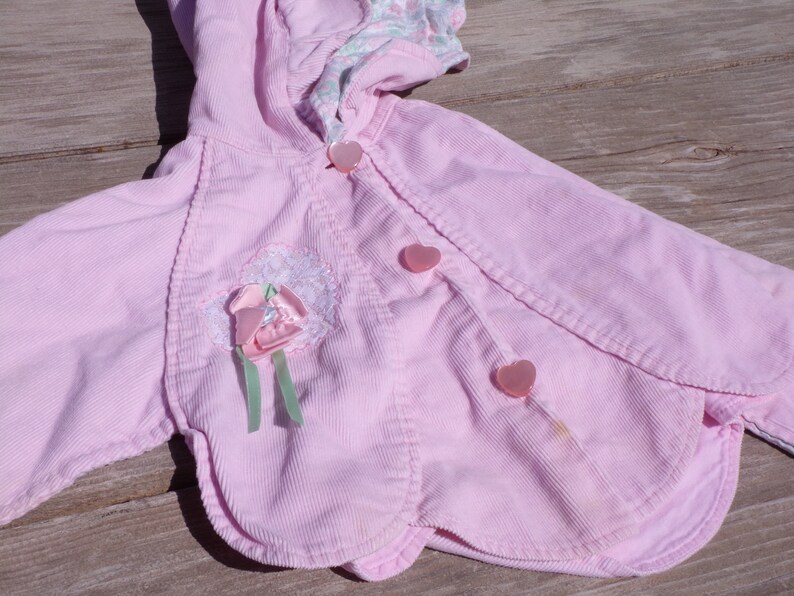 Puede incluir: Una chaqueta de pana rosa con capucha y botones en forma de coraz&oacute;n. La chaqueta tiene un adorno de flor en la parte delantera.