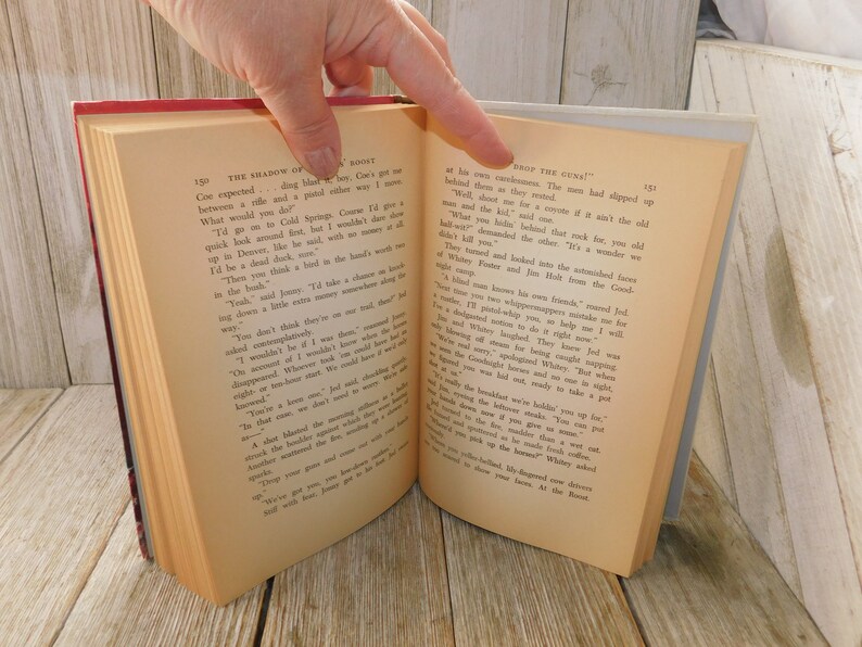 May include: Open book with a cream-colored cover and black text. The text on the left page reads "The Shadow of the Boint" and the text on the right page reads "Drop the Gun!"