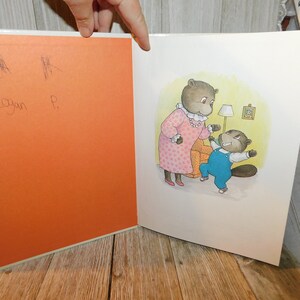 May include: A children's book illustration of two beavers, a mother and a child, in a living room setting. The mother beaver is wearing a pink dress with white polka dots and a pearl necklace. The child beaver is wearing blue overalls and a white shirt. The child beaver is holding a toy and looking at the mother beaver.
