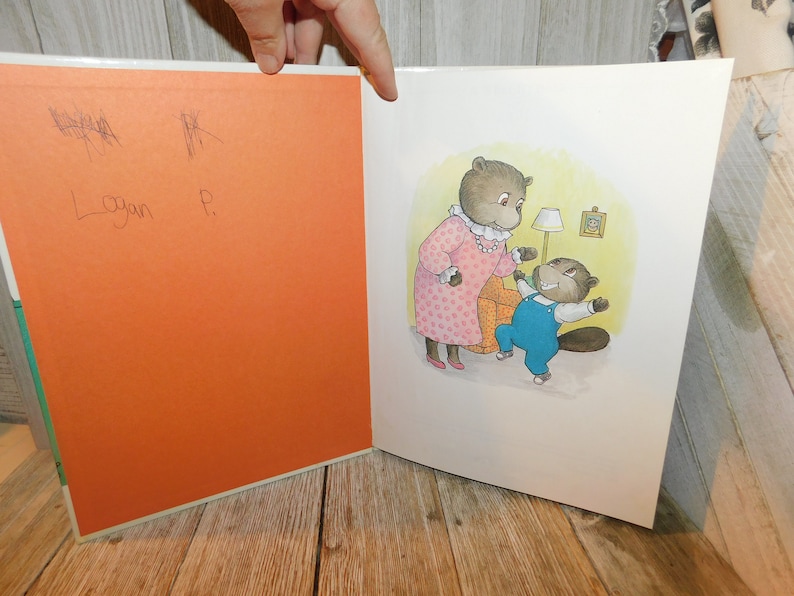 May include: A children's book illustration of two beavers, a mother and a child, in a living room setting. The mother beaver is wearing a pink dress with white polka dots and a pearl necklace. The child beaver is wearing blue overalls and a white shirt. The child beaver is holding a toy and looking at the mother beaver.