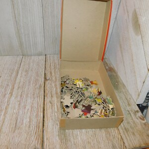 May include: A cardboard box filled with a colorful jigsaw puzzle. The pieces are scattered inside the box, which is open and sitting on a wooden surface.