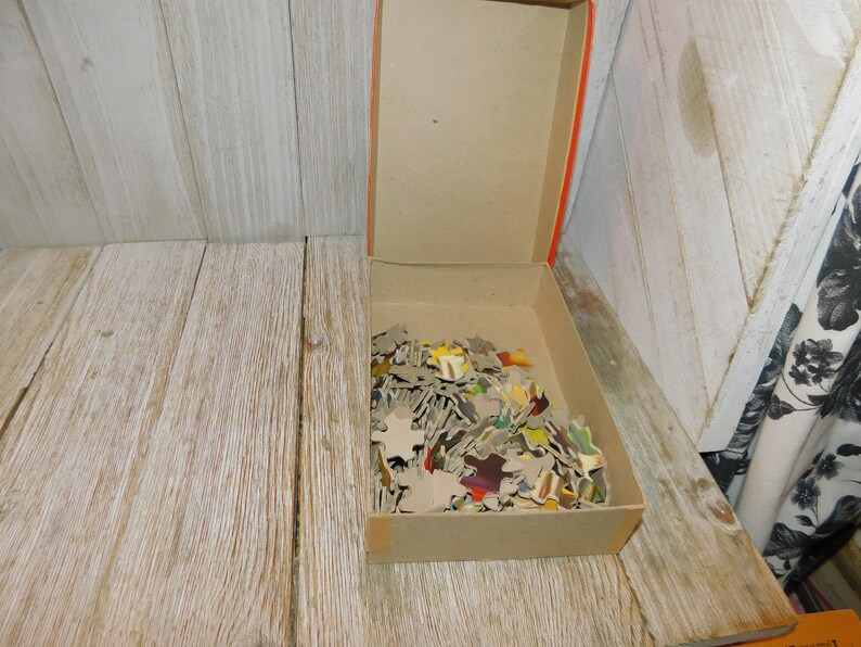 May include: A cardboard box filled with a colorful jigsaw puzzle. The pieces are scattered inside the box, which is open and sitting on a wooden surface.