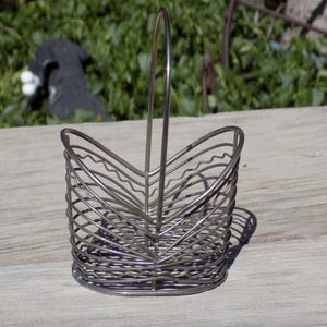 May include: A silver-colored wire basket with a handle. The basket has a rounded base and a series of curved wires forming the sides. The handle is a single, arched wire. The basket is sitting on a weathered wooden surface.