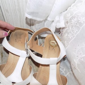 K&ouml;nnte beinhalten: Wei&szlig;e Ledersandalen mit T-Riemen. Die Sandalen haben eine braune Innensohle und der Markenname "b.o.c. comfort" ist auf der Innensohle aufgedruckt.