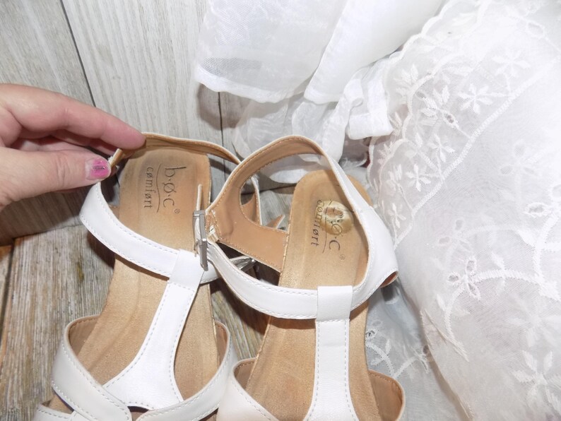 K&ouml;nnte beinhalten: Wei&szlig;e Ledersandalen mit T-Riemen. Die Sandalen haben eine braune Innensohle und der Markenname "b.o.c. comfort" ist auf der Innensohle aufgedruckt.