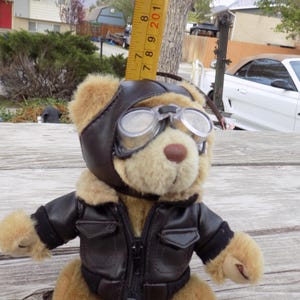 May include: A plush teddy bear wearing a brown leather aviator jacket, helmet, and goggles. The bear is tan with a brown nose and is posed on a wooden surface. A yellow ruler is placed next to the bear.