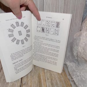 May include: A book open to two pages showing instructions for card games. The first page shows a diagram of a circle with 12 cards arranged around it. The second page shows a diagram of 9 cards arranged in a 3x3 grid. The text on the first page reads "Pirate Gold". The text on the second page reads "Solitaire".