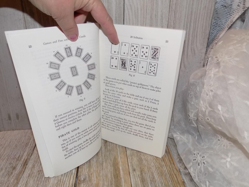 May include: A book open to two pages showing instructions for card games. The first page shows a diagram of a circle with 12 cards arranged around it. The second page shows a diagram of 9 cards arranged in a 3x3 grid. The text on the first page reads "Pirate Gold". The text on the second page reads "Solitaire".