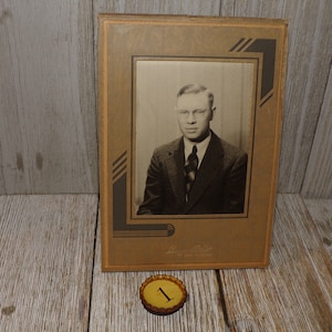 May include: A black and white photograph of a man wearing a suit and tie. The photo is framed in a brown frame with a decorative border. The frame has a gold inscription that reads "Lester Photo."