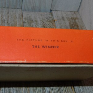 May include: Close-up of an orange box with the text "THE PICTURE IN THIS BOX IS THE WINNER" printed on it.