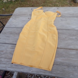 May include: A yellow sleeveless midi dress with a fitted bodice and a straight skirt. The dress has a sweetheart neckline and a zipper closure in the back.
