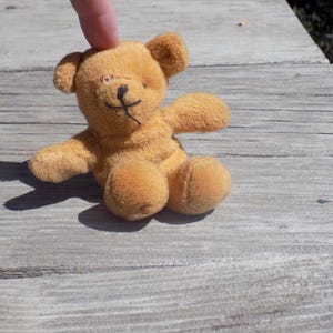 May include: A small, brown teddy bear with black eyes and a stitched nose. The bear is sitting on a wooden surface.