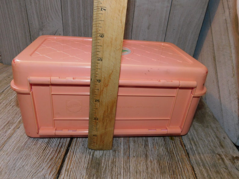 Puede incluir: Una caja de almacenamiento de pl&aacute;stico rosa con una tapa con bisagras y un cierre. La caja tiene una superficie texturizada y una peque&ntilde;a pegatina blanca en la parte superior.