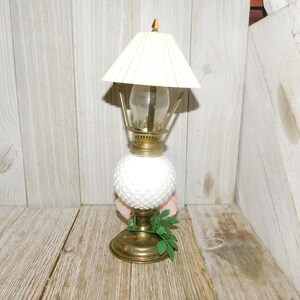 May include: A white milk glass oil lamp with a brass base and a white pleated lampshade. The lamp is sitting on a wooden surface.