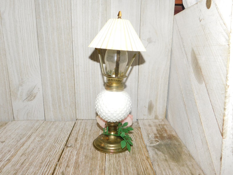 May include: A white milk glass oil lamp with a brass base and a white pleated lampshade. The lamp is sitting on a wooden surface.