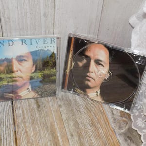 May include: Two CD cases are displayed. The first case has the text "WIND RIVER Vasquez" on the cover, with an image of a man and a landscape. The second case is open, revealing the CD with the same image.