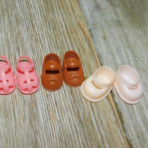 May include: A set of six doll shoes in pink, brown, and white. The shoes are made of plastic and have a simple design.