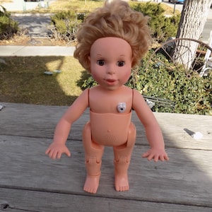 May include: A vintage baby doll with blonde, curly hair and brown eyes. The doll is wearing a peach-colored outfit and has a speaker on its chest. The doll is standing on a wooden surface.