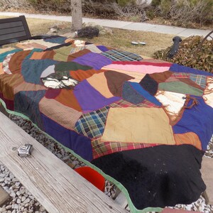 May include: A patchwork quilt made from various fabrics in shades of brown, blue, green, purple, and beige. The quilt is laid out on a wooden picnic table.