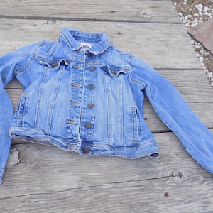 May include: A light blue denim jacket featuring a classic collar, long sleeves with buttoned cuffs, and a front button-down closure. The jacket includes two chest pockets with buttoned flaps and two vertical side pockets, finished with visible stitching.