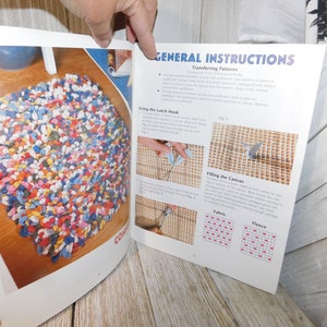 May include: A close-up of a book with instructions on how to use a latch hook to create a rug. The book shows a finished rug made of colorful fabric scraps, as well as diagrams of how to fill the canvas with fabric or fleece.