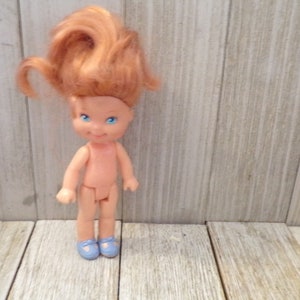 Pode incluir: Uma pequena boneca nua com cabelo ruivo e olhos azuis. A boneca est&aacute; usando sapatos azuis e est&aacute; em p&eacute; em uma superf&iacute;cie de madeira.