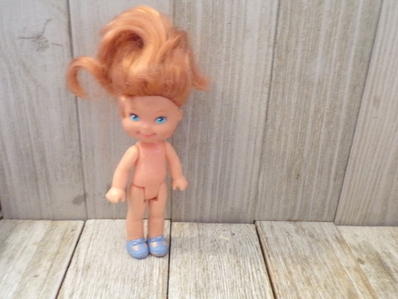 Pode incluir: Uma pequena boneca nua com cabelo ruivo e olhos azuis. A boneca est&aacute; usando sapatos azuis e est&aacute; em p&eacute; em uma superf&iacute;cie de madeira.
