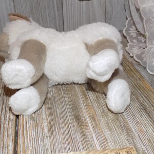 May include: A plush toy cat in a lying position, featuring a soft, white body with tan patches on its paws, ears, and sides. The toy is made of a fluffy material, giving it a cuddly appearance. The cat is lying on a wooden surface.