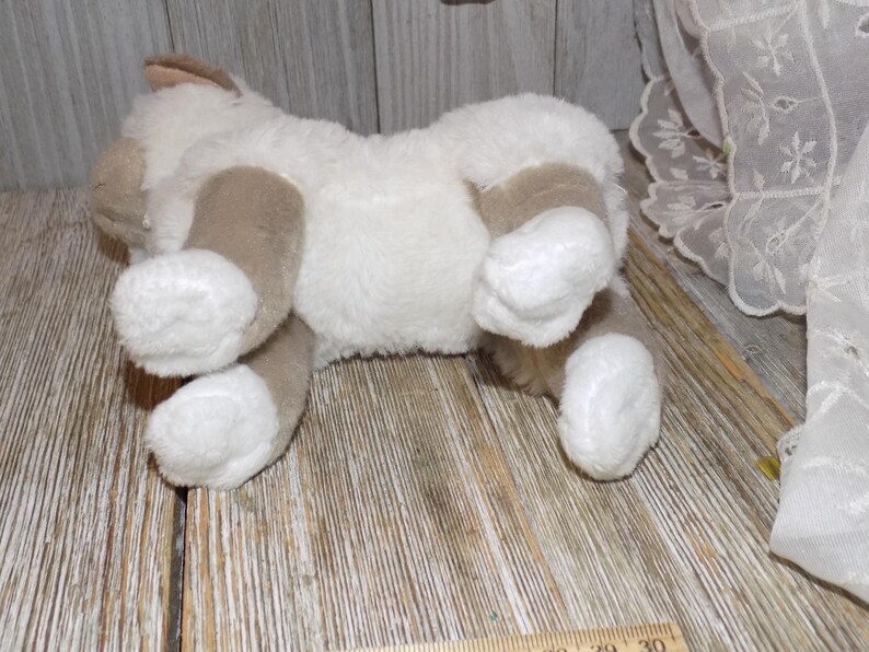 May include: A plush toy cat in a lying position, featuring a soft, white body with tan patches on its paws, ears, and sides. The toy is made of a fluffy material, giving it a cuddly appearance. The cat is lying on a wooden surface.