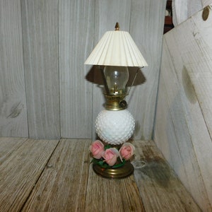 May include: A white milk glass oil lamp with a white fabric lampshade and three pink roses on the base. The lamp is on a wooden surface.
