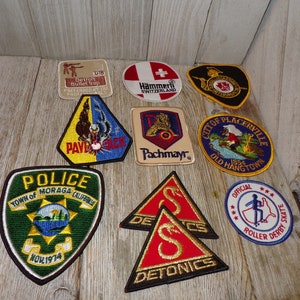 May include: A collection of nine embroidered patches featuring various designs, including a police badge, a roller derby logo, a city seal, and a gun manufacturer logo. The patches are all different colors and sizes and are arranged in a random pattern on a wooden surface.