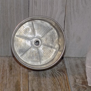 May include: A silver metal coffee pot filter with a circular shape and a perforated design. The filter has a central hole and multiple radiating lines.