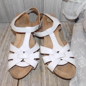 K&ouml;nnte beinhalten: Wei&szlig;e Ledersandalen mit Korkkeil und T-Riemen. Die Sandalen haben ein Kreuzmuster an der Vorderseite und einen Schnallenverschluss am Kn&ouml;chelriemen.