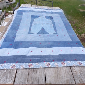May include: A handmade denim quilt with a patchwork design. The quilt features various shades of blue denim, with red stitching accents and a light blue border with a teddy bear pattern. A pair of denim overalls is sewn into the center.