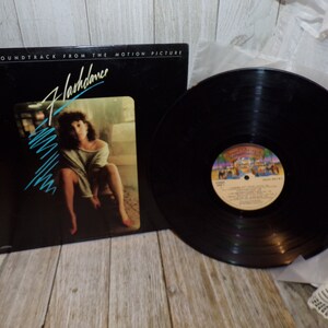 May include: A black vinyl record with a white label featuring the text "Flashdance" and a colorful image of a beach scene. The record is on a white surface with a white and black patterned fabric in the background. The album cover is black with a photo of a woman in a white shirt and a blue and white striped background.