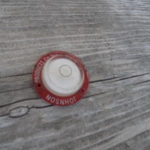 May include: A circular Johnson Products Co. Inc. bullseye spirit level featuring a red metal base with white text and a clear, domed liquid-filled center with a black circular target line for indicating horizontal surface alignment.