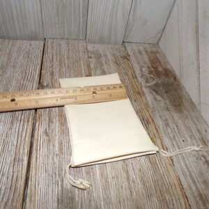 Puede incluir: Una peque&ntilde;a bolsa de cord&oacute;n de color blanco roto hecha de tela. La bolsa est&aacute; sobre una superficie de madera con una regla al lado. La regla mide 76 cm de largo.