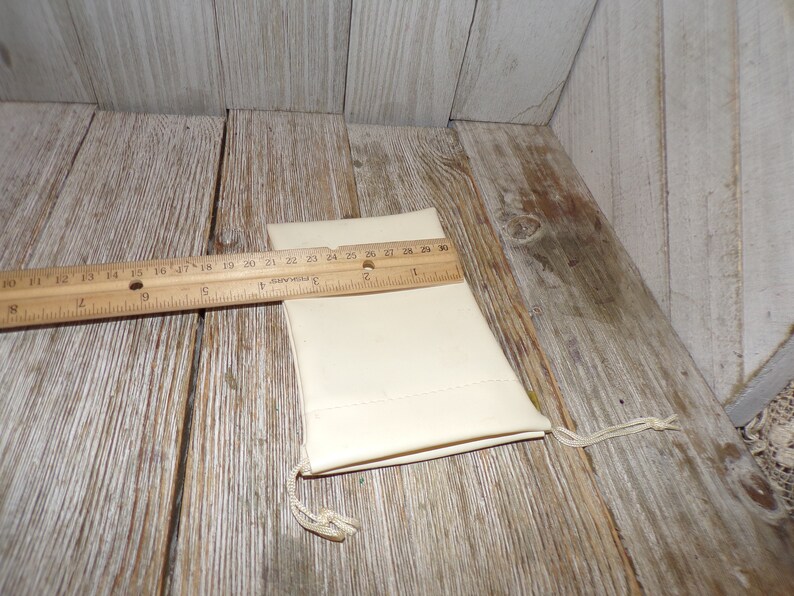 Puede incluir: Una peque&ntilde;a bolsa de cord&oacute;n de color blanco roto hecha de tela. La bolsa est&aacute; sobre una superficie de madera con una regla al lado. La regla mide 76 cm de largo.