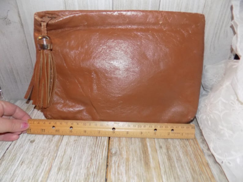 May include: A brown leather clutch with a tassel and a metal accent. The clutch is about 10 inches long.
