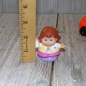 May include: A small plastic doll with brown hair, wearing a yellow jacket, pink skirt, and purple shoes. The doll is holding a white bottle in one hand and has a pink heart on the other side of the jacket.