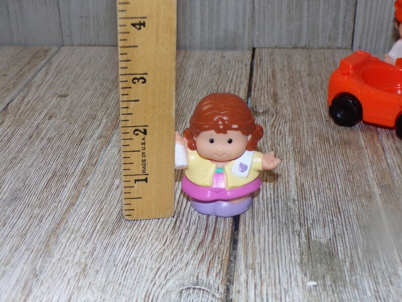 May include: A small plastic doll with brown hair, wearing a yellow jacket, pink skirt, and purple shoes. The doll is holding a white bottle in one hand and has a pink heart on the other side of the jacket.