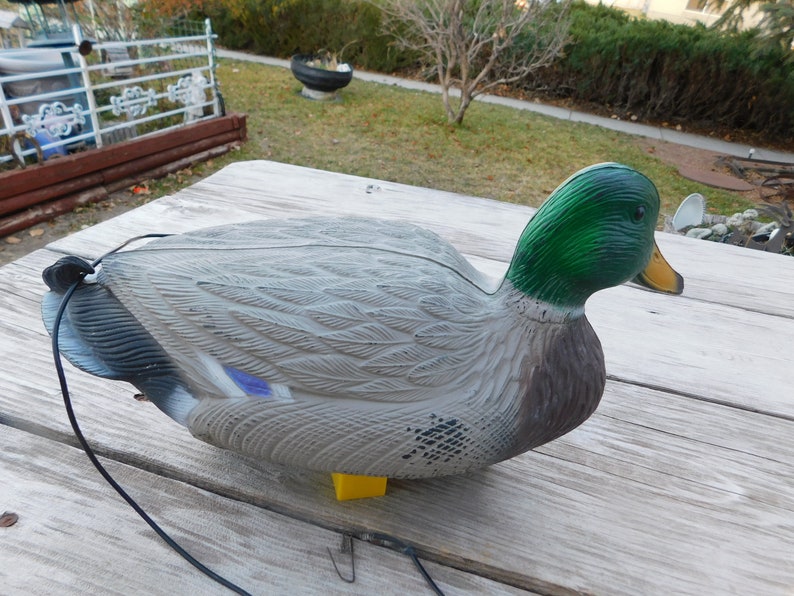 May include: A gray and brown plastic duck decoy with a green head and a yellow beak. The decoy has a black cord attached to it.
