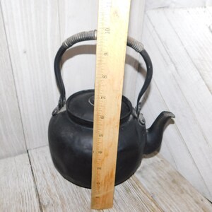 May include: Black cast iron teapot with a wire handle. The teapot is approximately 18 cm tall.