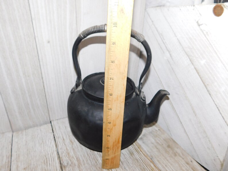May include: Black cast iron teapot with a wire handle. The teapot is approximately 18 cm tall.